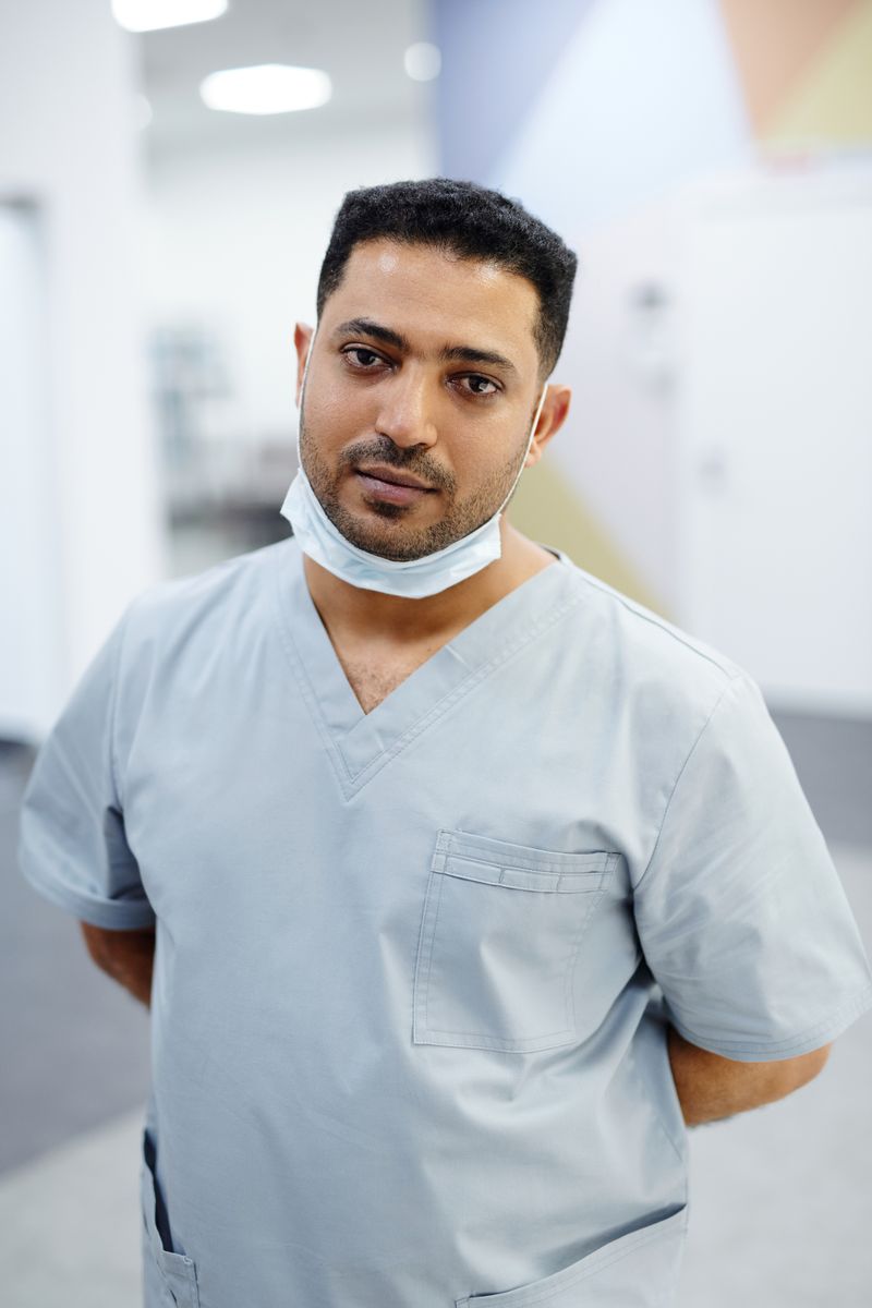 Contact A healthcare worker in blue scrubs standing indoors, with a mask around the neck.